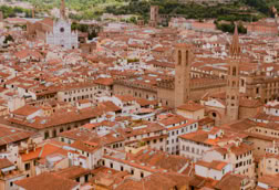 Aerial view of a historic city with red-tiled roofs and several towers, surrounded by green hills under a cloudy sky.